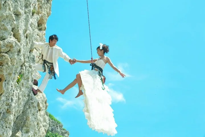 Rock Climbing wedding couple kissing while suspended on rope next to tan overhanging cliff.
